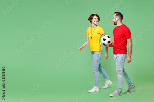 Full length of cheerful young couple friends sport family woman man football fans in t-shirts cheer up support favorite team with soccer ball walking going looking aside isolated on green background.