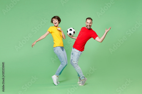 Full length of laughing couple friends sport family woman man football fans in t-shirts cheer up support favorite team with soccer ball standing on toes spreading hands isolated on green background.