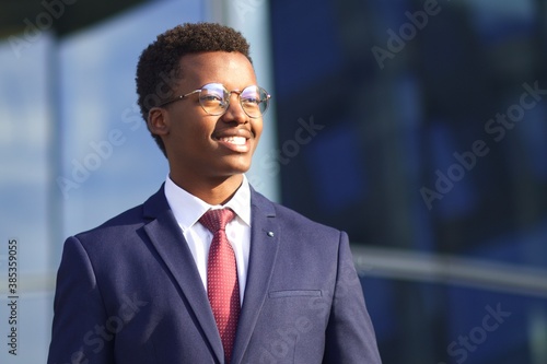 Portrait of handsome happy cheerful positive businessman in formal suit, tie and glasses. Black African Afro American successful man standing outdoors business building, smiling, looking into distance