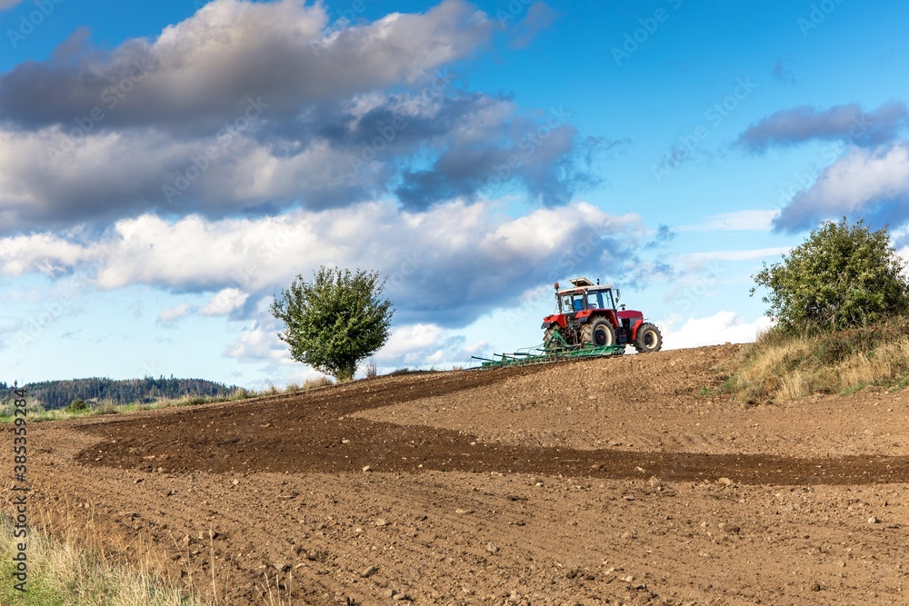 Obraz premium Red tractor cultivates the soil in the field with a cultivator after harvest. Autumn sunny day. Agricultural landscape in the Czech Republic. Work on the farm.