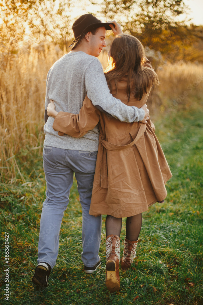Fototapeta premium Happy stylish couple hugging and walking in autumn meadow in warm sunset light
