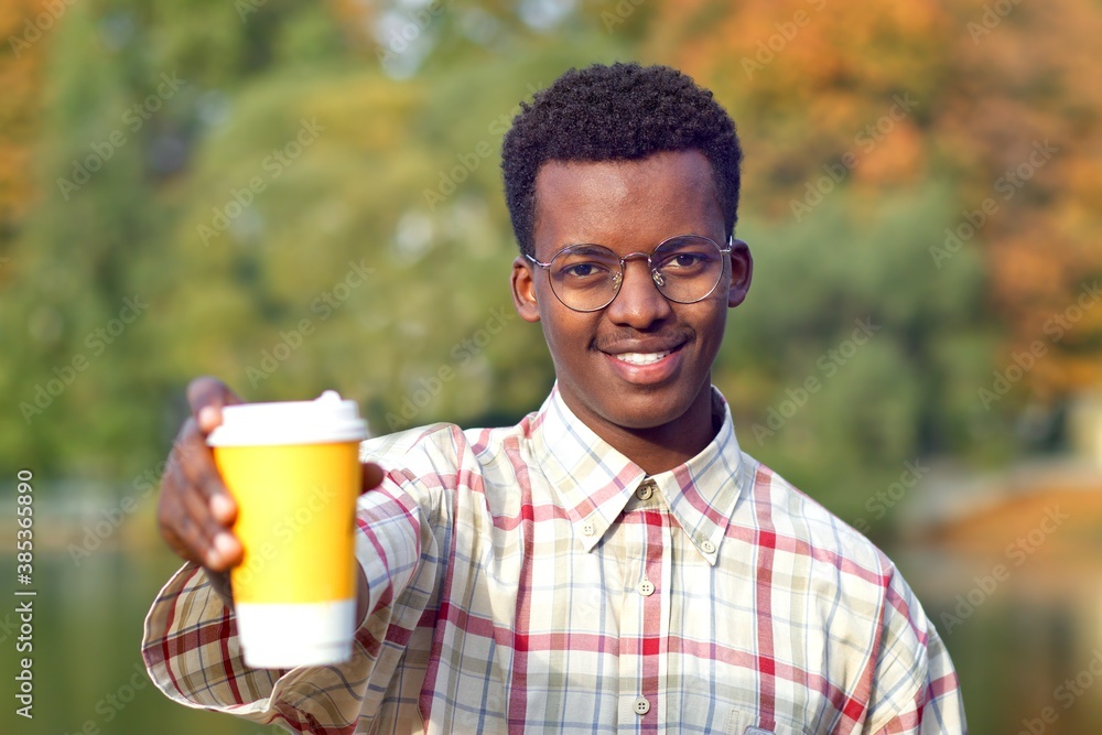 Portrait of young happy positive man in shirt and glasses holding out a plastic cup of hot drink tea or coffee, smiling, looking at camera. Black African Afro American guy in golden autumn park. Portrait of young happy positive man in shirt and glasses holding out a plastic cup of hot drink tea or coffee, smiling, looking at camera. Black African Afro American guy in golden autumn park.
