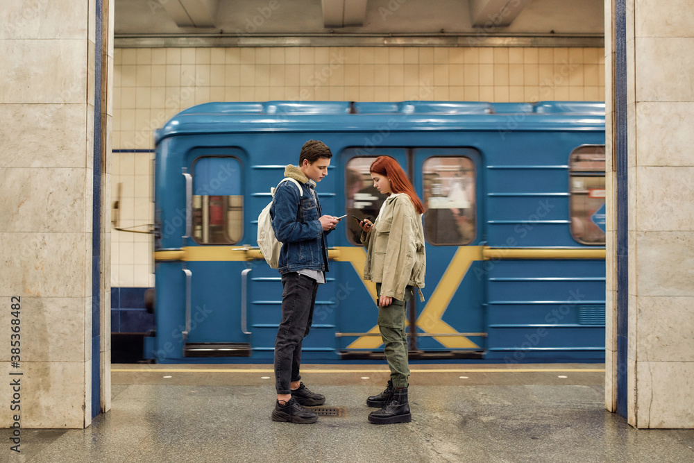 Full length shot of couple of teenagers using smartphones, totally absorbed in online life, ignoring each other while standing at the subway metro station