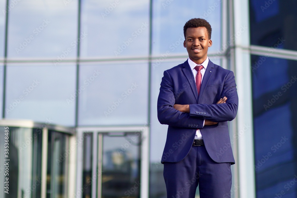 Fototapeta premium Portrait of serious confident successful black African man, Afro American young businessman in formal suit with tie standing outdoors business office with his hands crossed, looking at camera