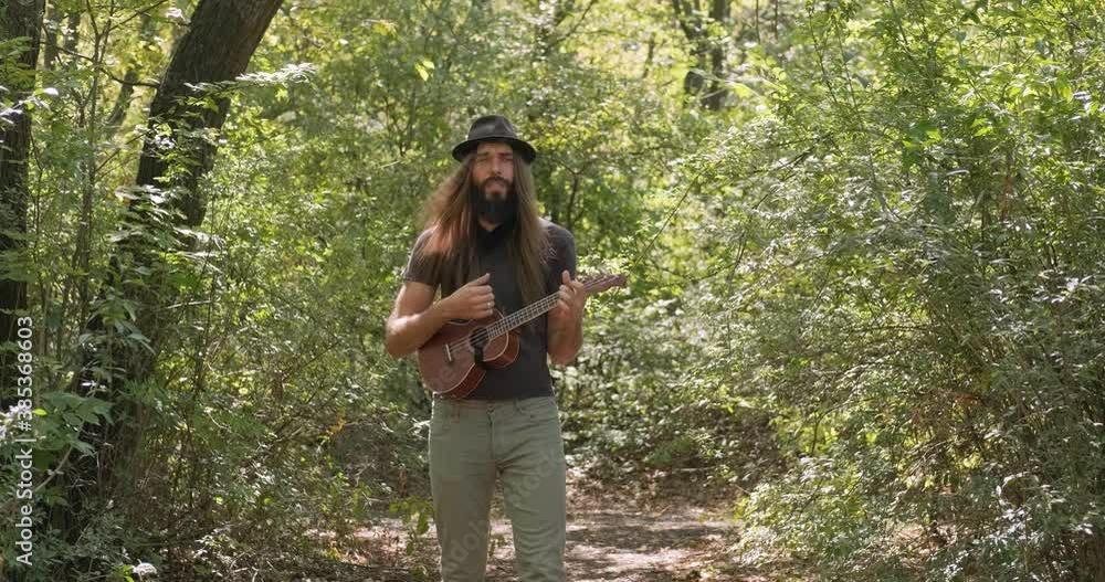 Bearded male with longhait and hat posing with ukulele in the forest Bearded male with longhait and hat posing with ukulele in the forest