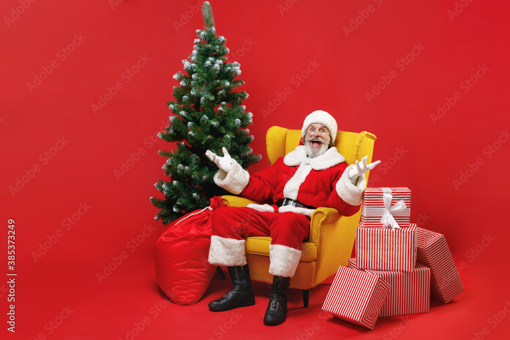 Surprised Santa Claus man in Christmas hat suit coat sit in armchair with fir tree presents gifts spreading hands isolated on red background studio. Happy New Year celebration merry holiday concept. Surprised Santa Claus man in Christmas hat suit coat sit in armchair with fir tree presents gifts spreading hands isolated on red background studio. Happy New Year celebration merry holiday concept.