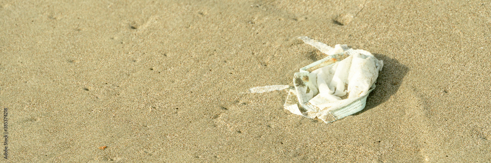 White face medical mask is left abandoned on sand beach. Garbage polluting the natural environment. Wide banner White face medical mask is left abandoned on sand beach. Garbage polluting the natural environment. Wide banner