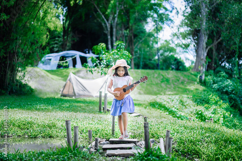 little asian girl playing ukulele or hawaiian guitar in the park while camping in summertime