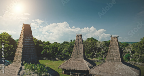 Sun light at high straw roof of traditional house in Kodi village, Indonesia. Aerial view of primitive Indonesian settlement admist green tropic forest. Amazing summer vacation at unique Asia landmark photo