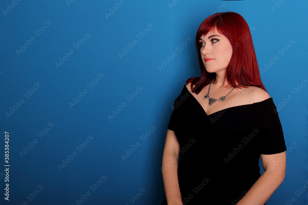 Naklejka premium Young woman looking aside, sad sadness look, red haired girl in black dress thinking face on blue background, studio shot, copy space