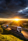 Trucks and delivery vans driving on the asphalt highway in forested landscape in the golden rays of the sunset with dark cloud