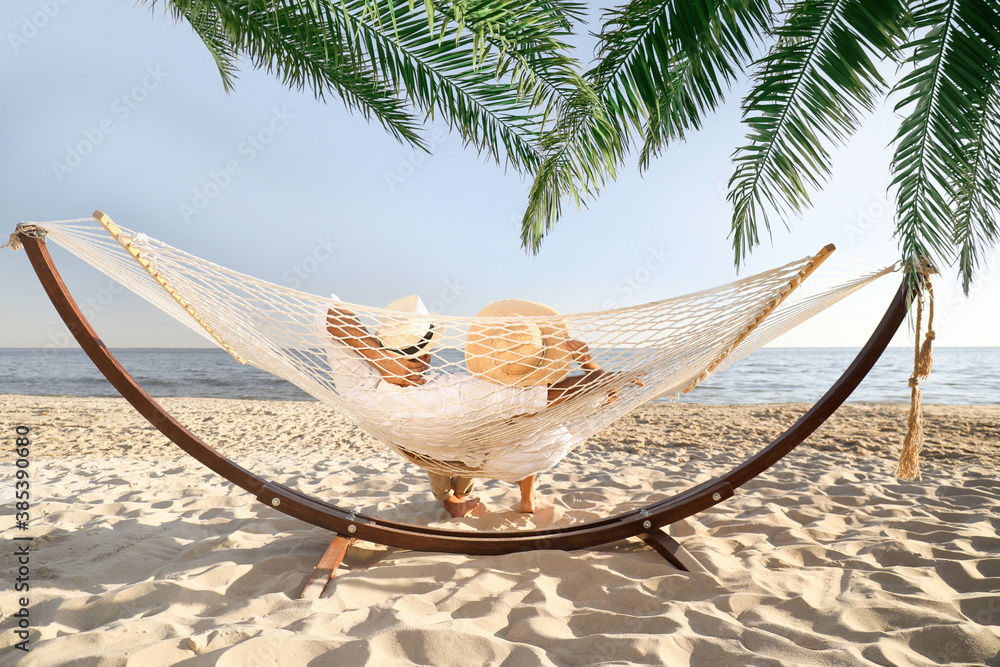 Couple relaxing in hammock under green palm leaves on beach - obrazy, fototapety, plakaty 