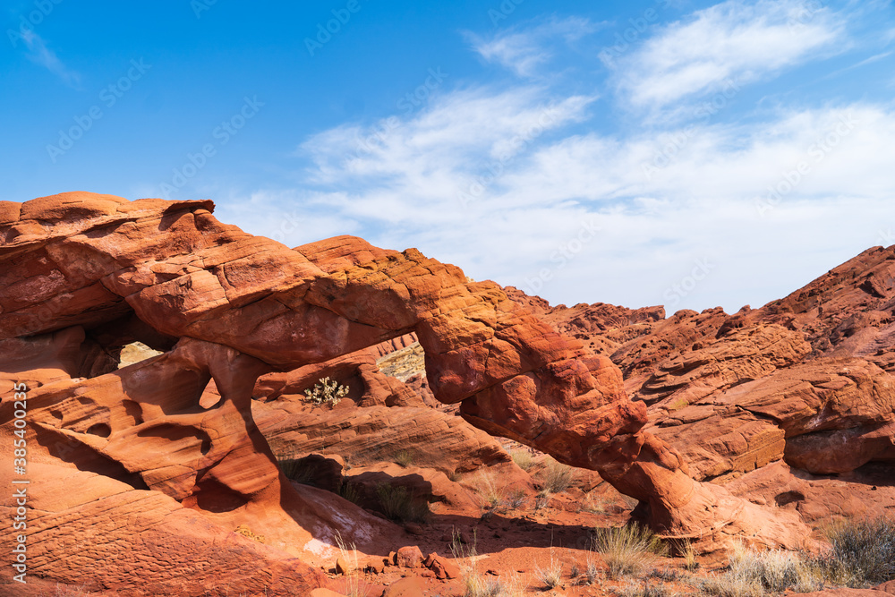 Fototapeta premium A natural sandstone arch in a red desert landscape under a clear blue sky with scattered clouds.