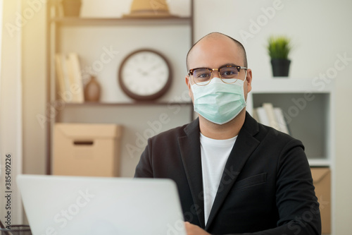 Young businessman wearing protection mask working with laptop in office. Social distancing safety from corona virus flu.