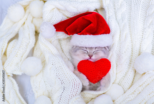 A small light tabby kitten lies on its back on a white knitted scarf in a Santa hat and holds a plush heart with its front paws