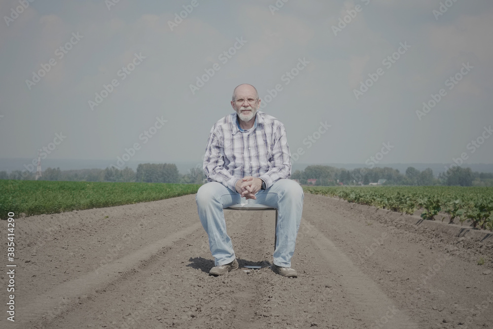 An elderly single man sits on a chair in the middle of a field and looking at the camera