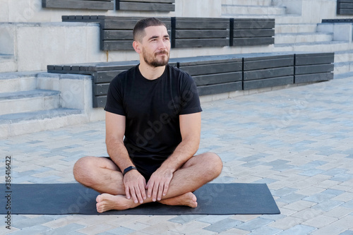 Man athlete in black sportswear practices yoga exercises in lotus position outdoors.
