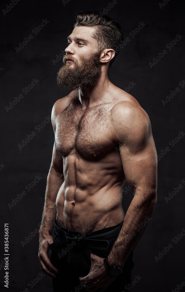 Cool and haired shirtless man with beard and moustache in pants posing with hands in pockets in dark background.
