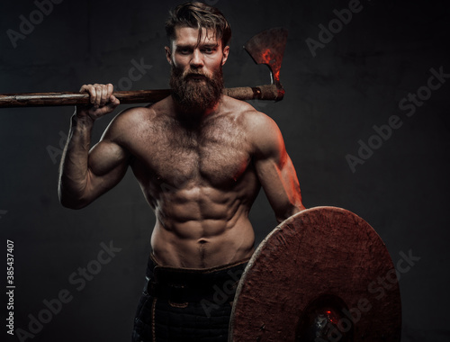 Foto Warlike and furious viking with naked torso and beard staying in dark background holding shield and two handed axe behind his back