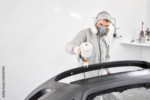 Worker painting parts of the car in special painting chamber, wearing costume and protective gear. Car service station.