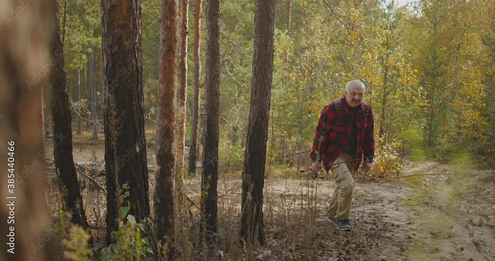 grey-haired man with spin fishing rod in hand is walking to shore of river through forest area during travel