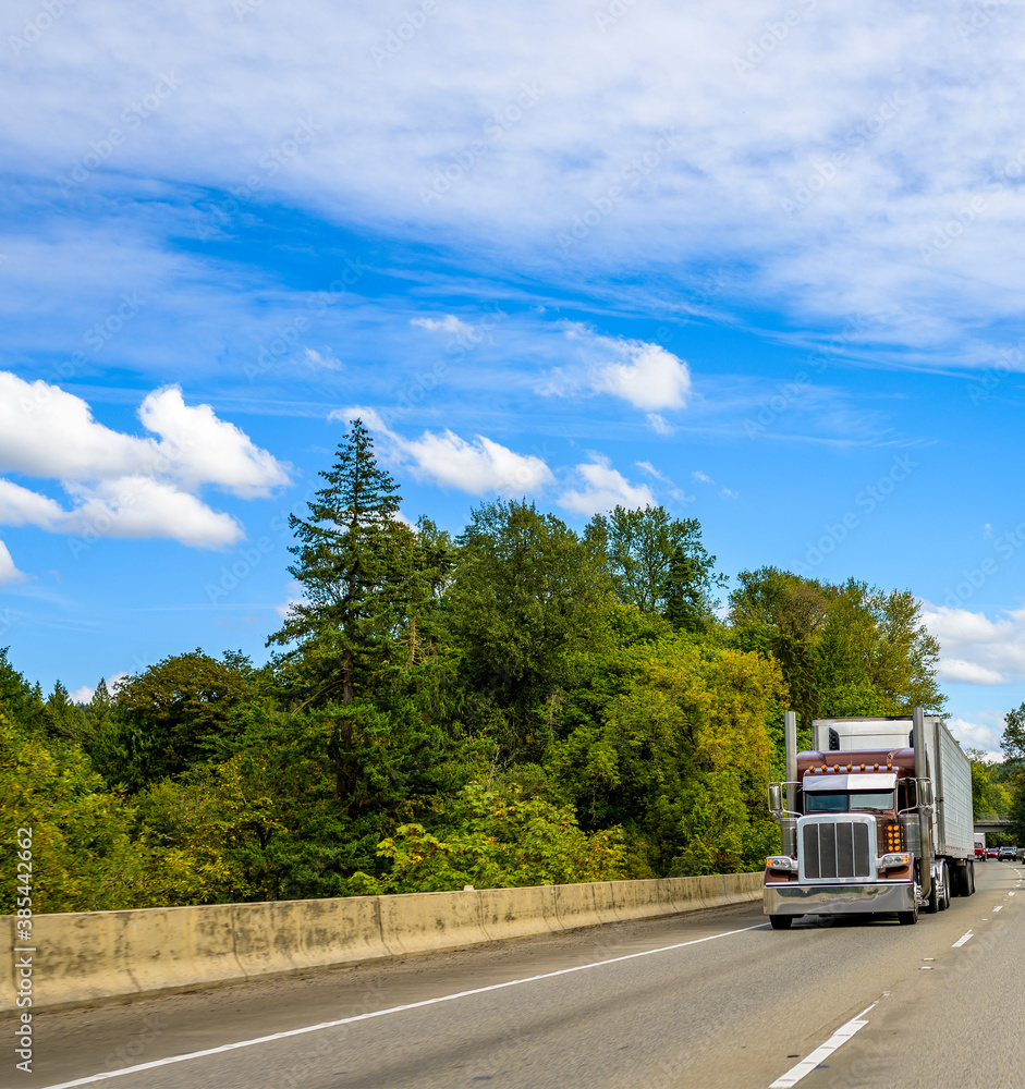 Big rig classic burgundy semi truck with lights transporting cargo in refrigerator semi trailer driving on the straight highway road Big rig classic burgundy semi truck with lights transporting cargo in refrigerator semi trailer driving on the straight highway road