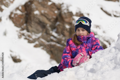 beautiful happy little girl in ski suit and wool hat with goggles lying on white snow in mountains during winter outdoor leisure christmas vacation with copy space
