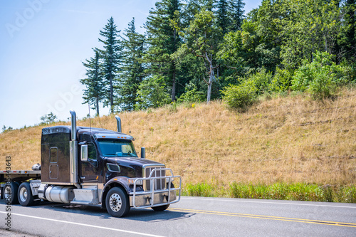 Wallpaper Mural Classic dark big rig semi truck with large grille guard and low cab for better airflow transporting flat bed semi trailer running on the road with hills with trees on the side Torontodigital.ca