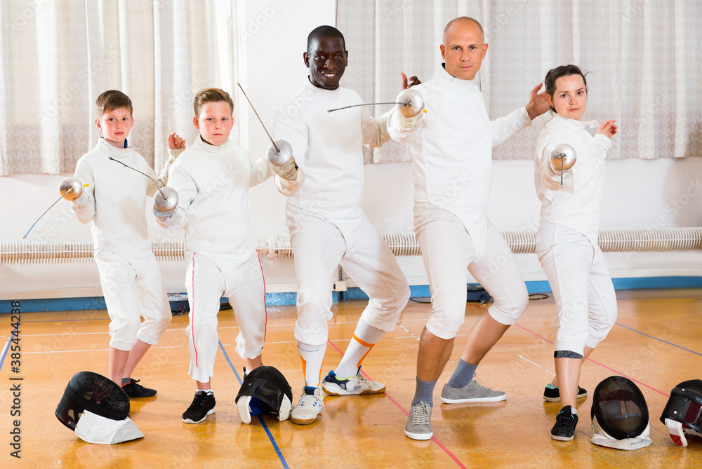 Group portrait of young happy cheerful fencers with coaches holding rapiers in training room