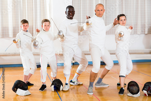 Group portrait of young happy cheerful fencers with coaches holding rapiers in training room