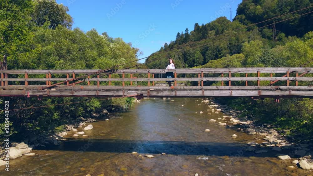 Aerial view from Old wooden suspension bridge over a mountain river. Girl standing on a wooden bridge and looking to the future. Punitive landscape of a mountain river on a sunny day. Aerial view from Old wooden suspension bridge over a mountain river. Girl standing on a wooden bridge and looking to the future. Punitive landscape of a mountain river on a sunny day.