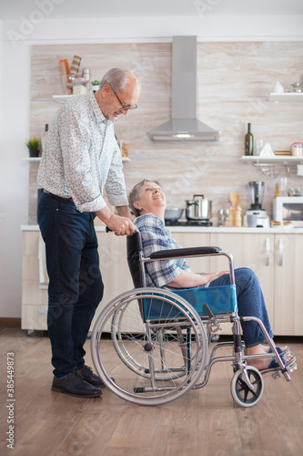 Husband looking at disabled senior woman in the kitchen. Disabled senior woman sitting in wheelchair in kitchen looking through window. Living with handicapped person. Husband helping wife with