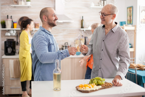 Happy father and son clinking wine glasses in kitchen during lunch with family.