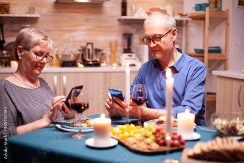 Browsing on smartphone in during their relationship anniversary in kitchen with candles on table. Sitting at the table in the kitchen, browsing, searching, using phone, internet, in the dining room.