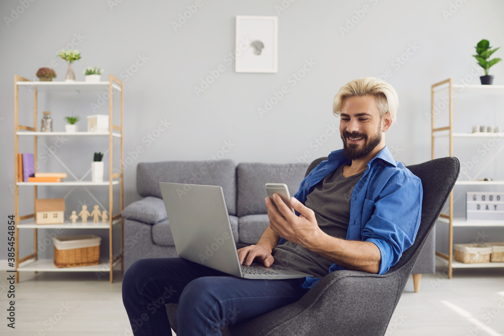 Successful freelancer is smiling while sitting in a chair with a laptop and mobile in hand texting message in the room.
