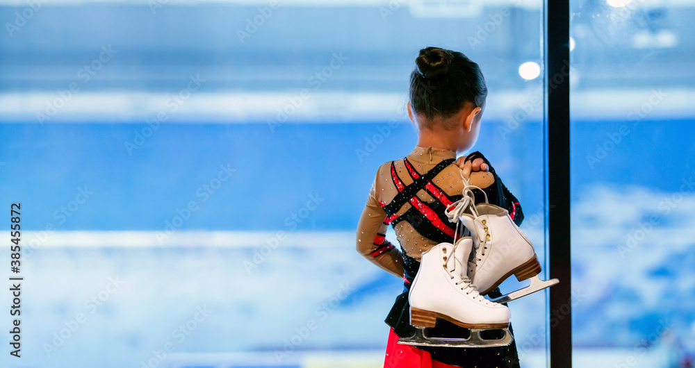 Fototapeta premium Back view portrait of little girl holding figure skates standing by ice rink and watching training, copy space