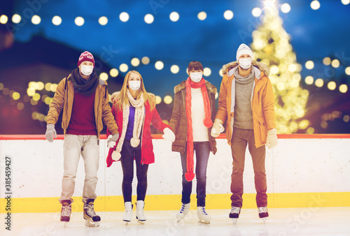 friendship, christmas and leisure concept - friends wearing face protective medical masks for protection from virus disease holding hands at outdoor skating rink over holiday lights background