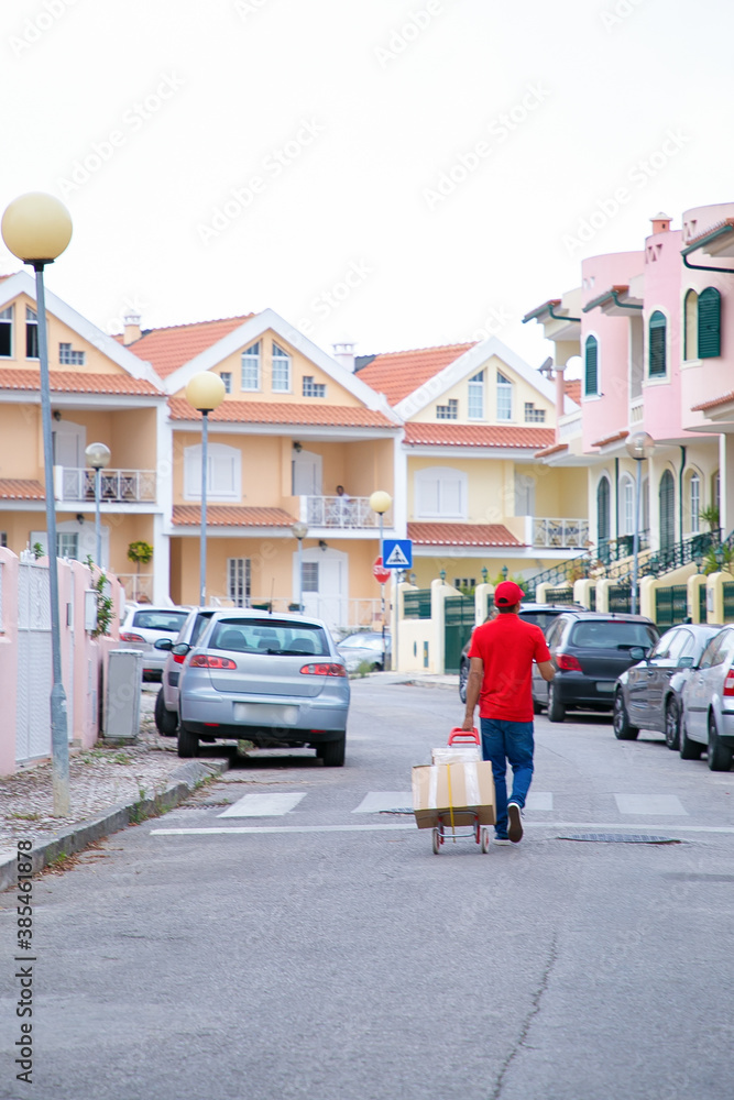 Postman walking in middle of street and wheeling trolley with boxes. Professional courier in red shirt a walking with cardboard parcels on cart. Houses on background. Delivery service and post concept