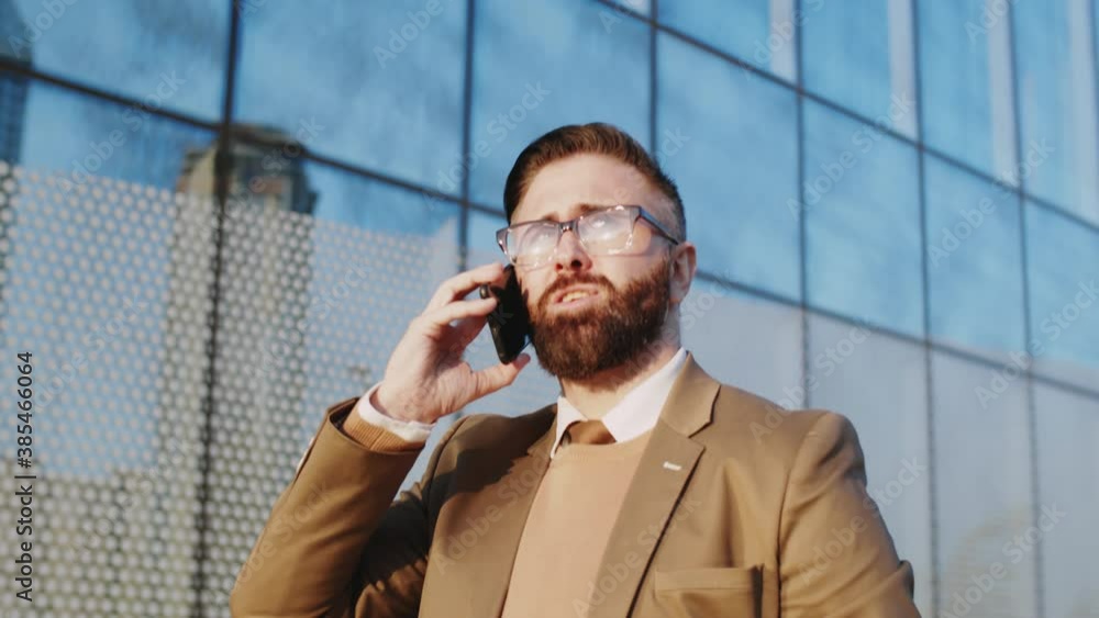 Low angle chest up shot of handsome bearded businessman in glasses and formal suit walking outdoors along modern office building and having phone talk Low angle chest up shot of handsome bearded businessman in glasses and formal suit walking outdoors along modern office building and having phone talk