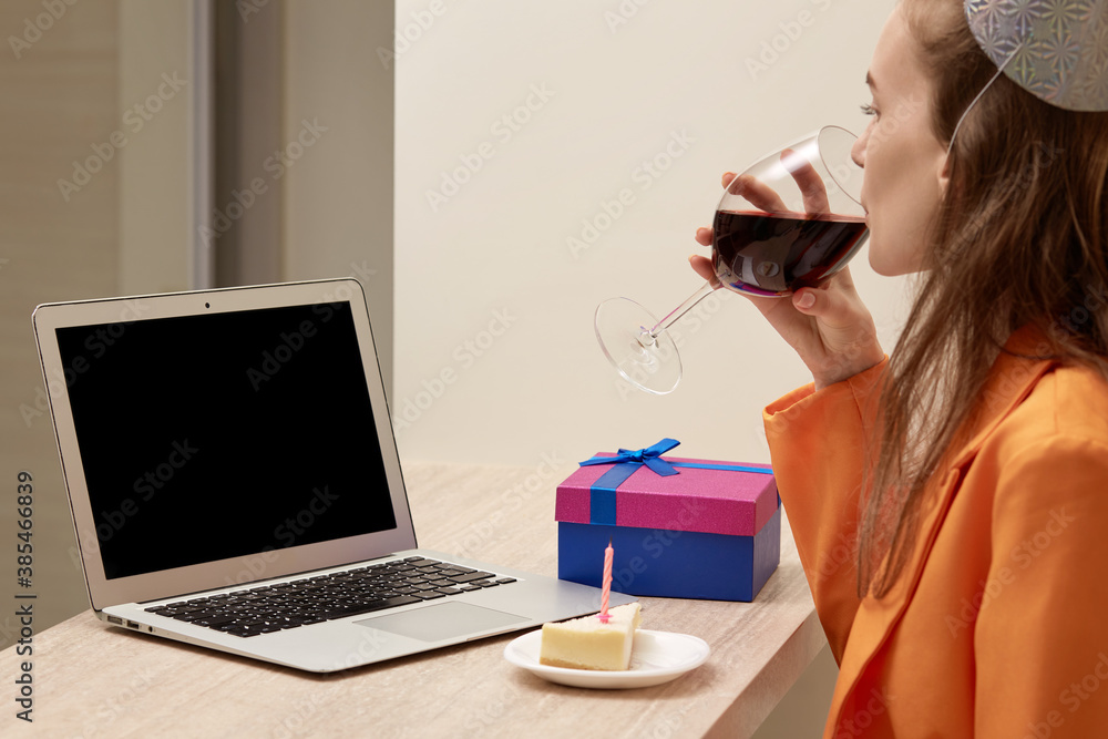 A young beautiful woman communicates online on her birthday with a glass of wine in her hands. On the table is a laptop with a black screen. Birthday celebrations during the pandemic. A young beautiful woman communicates online on her birthday with a glass of wine in her hands. On the table is a laptop with a black screen. Birthday celebrations during the pandemic.