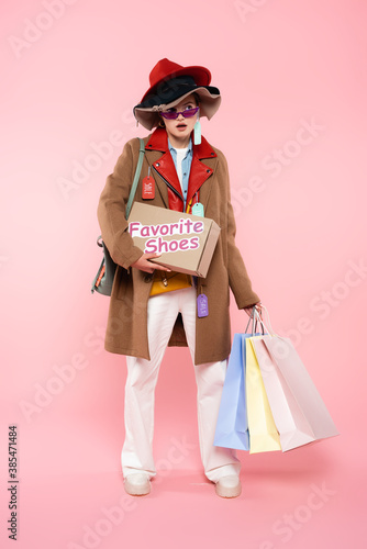 surprised woman in sunglasses and hats with sale tags holding box with favorite shoes lettering and shopping bags on pink, black friday concept