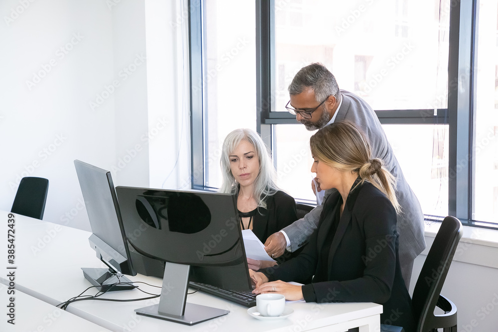 Serious business group of three analyzing reports, sitting at workplace with monitors together, holding, reviewing and discussing papers. Copy space. Business meeting concept Serious business group of three analyzing reports, sitting at workplace with monitors together, holding, reviewing and discussing papers. Copy space. Business meeting concept