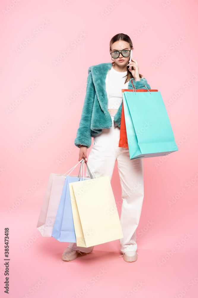 trendy woman in sunglasses talking on smartphone near shopping bags on pink, black friday concept