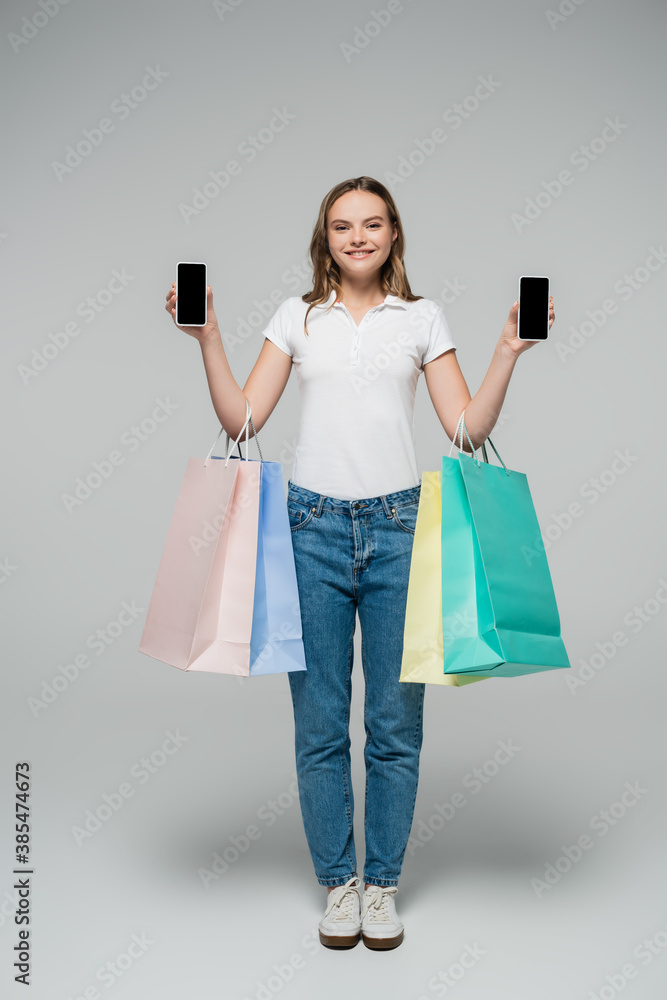 young joyful woman holding mobile phones with blank screen and shopping bags on grey, black friday concept young joyful woman holding mobile phones with blank screen and shopping bags on grey, black friday concept