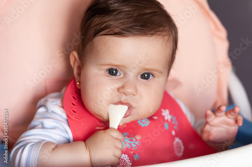 Mother gives baby food from a spoon. 6 month old baby girl eating porridge on highchair at kitchen.