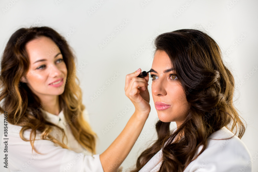 makeup artist coloring eyebrows with mascara to elegance woman with brown hair in white jacket at the white wall background makeup artist coloring eyebrows with mascara to elegance woman with brown hair in white jacket at the white wall background