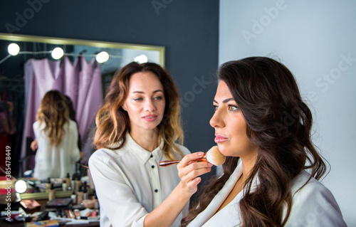 makeup artist makes a makeup with powder brush to elegance woman with brown hair in white jacket