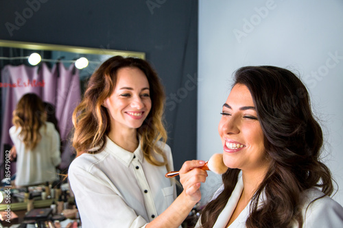 makeup artist makes a makeup with powder brush to elegance woman with brown hair in white jacket