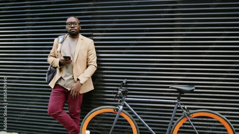 Zoom in shot of young stylish Afro-American businessman with backpack standing by bike outdoors, using smartphone and then posing for camera with confidence Zoom in shot of young stylish Afro-American businessman with backpack standing by bike outdoors, using smartphone and then posing for camera with confidence