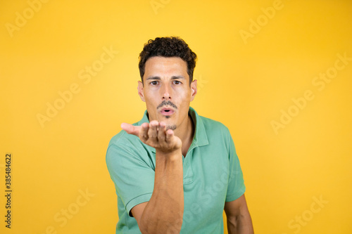 Young handsome man wearing green casual t-shirt over isolated yellow background looking at the camera blowing a kiss with hand on air being lovely and sexy. Love expression.